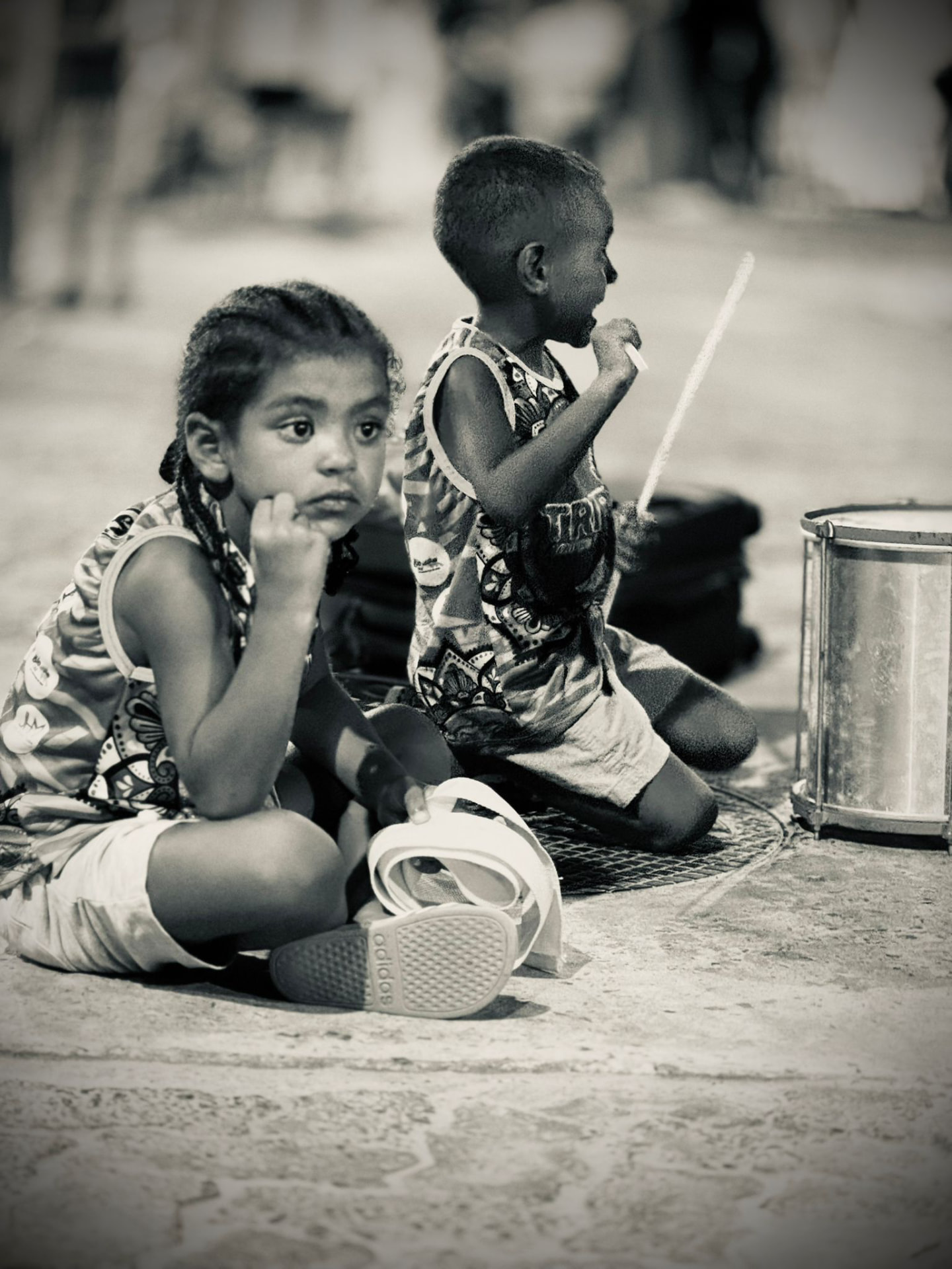 Two small children in TRIBO vests sitting on the ground beside a caixa drum watching a rehearsal