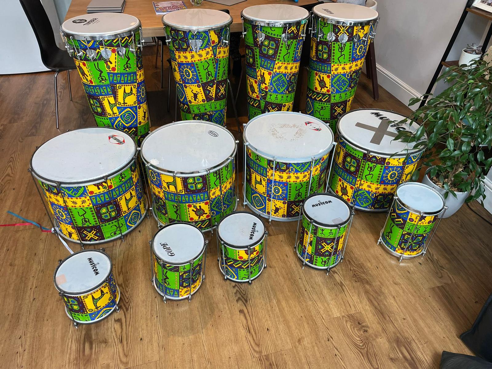 Set of hand-painted Brazilian percussion instruments — surdos, repiniques and timbaus — arranged on a wooden floor