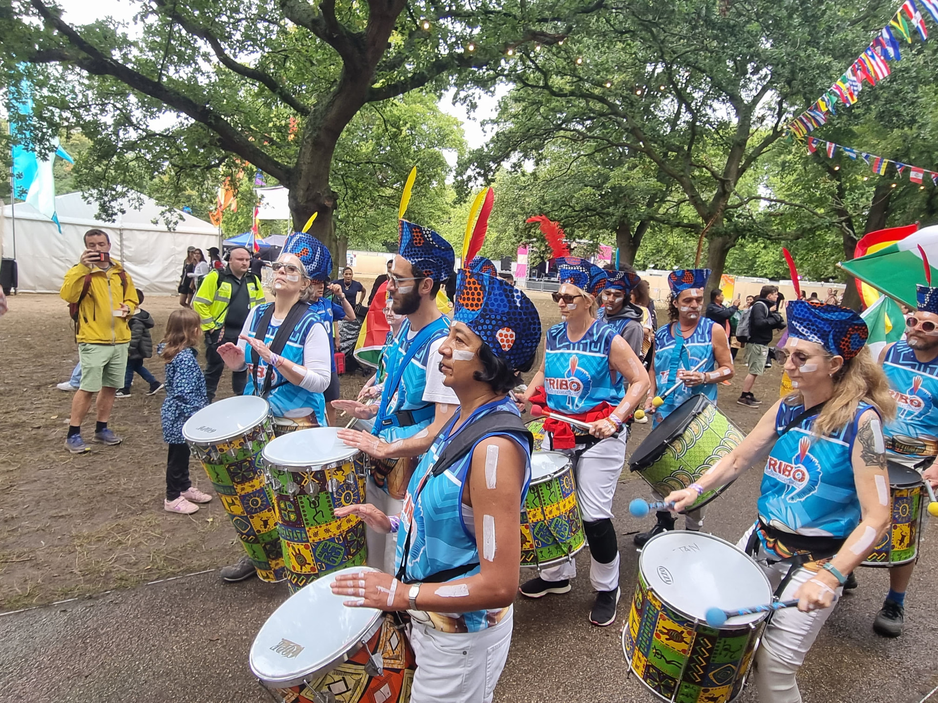 TRIBO percussionists parading through a London park with painted surdos and blue feathered headdresses