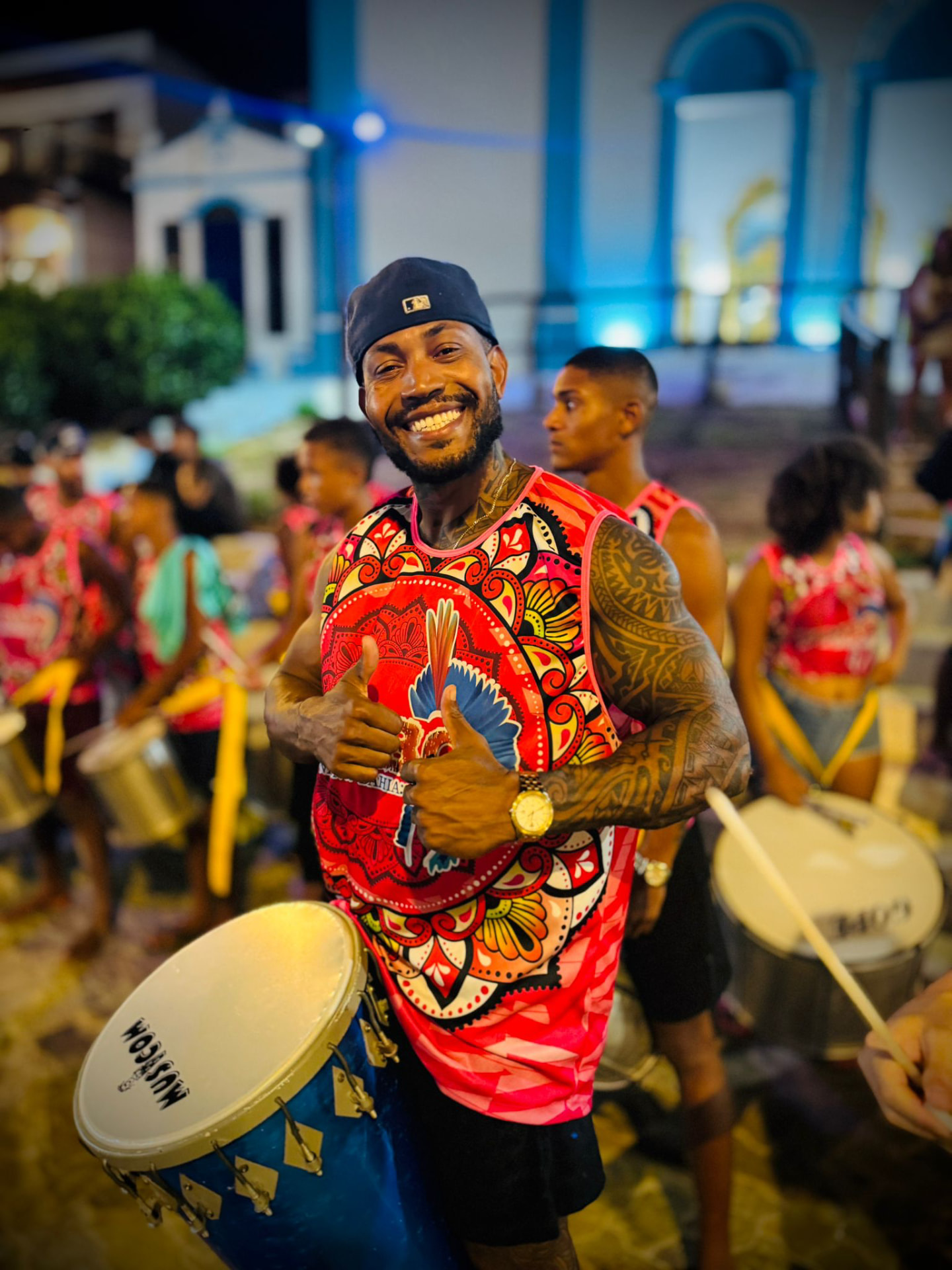 Smiling TRIBO drummer with a surdo giving a thumbs up in front of a blue-lit church at night