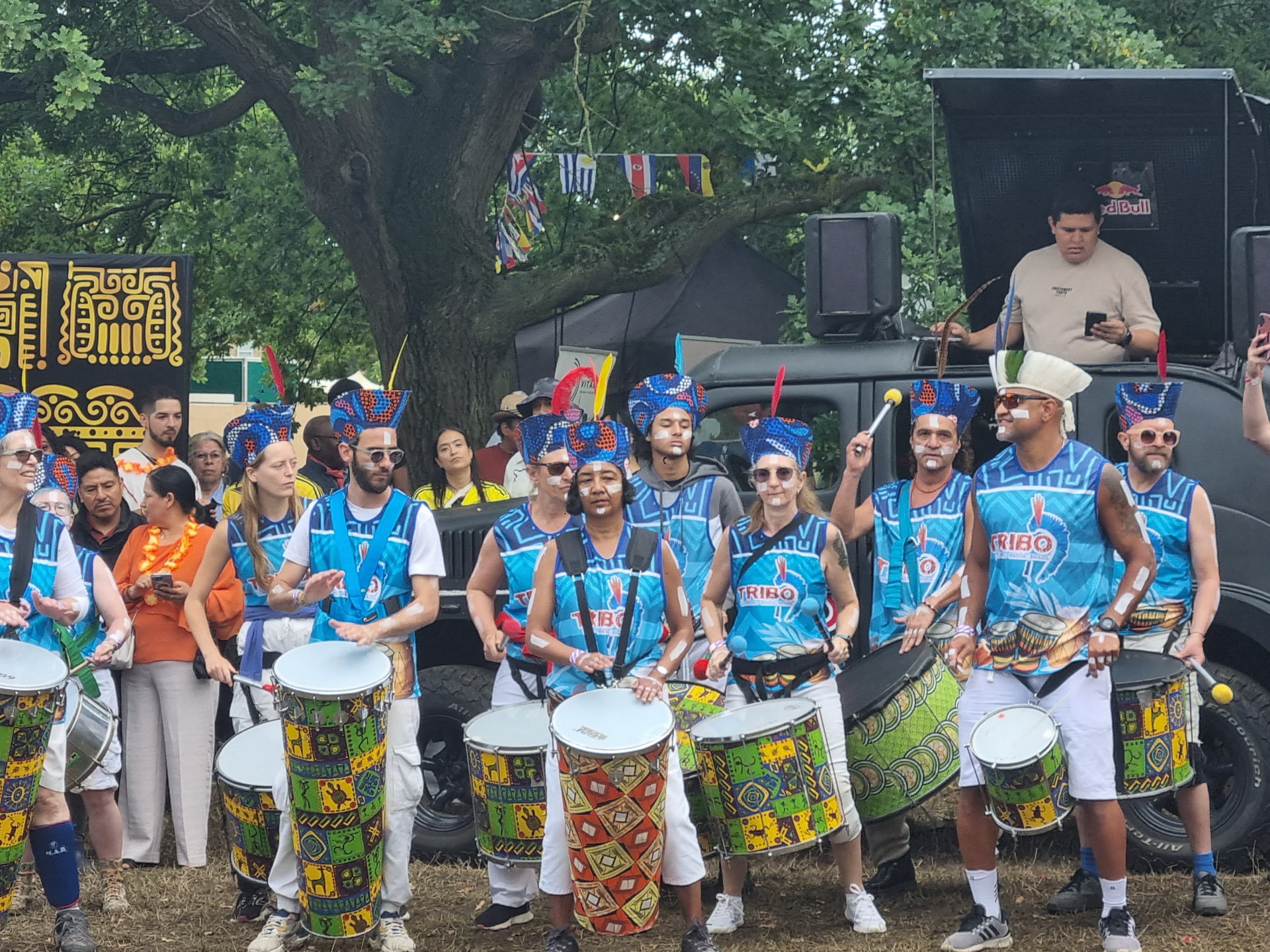 TRIBO drummers in blue band vests and feathered headdresses lined up with painted surdos at a London festival