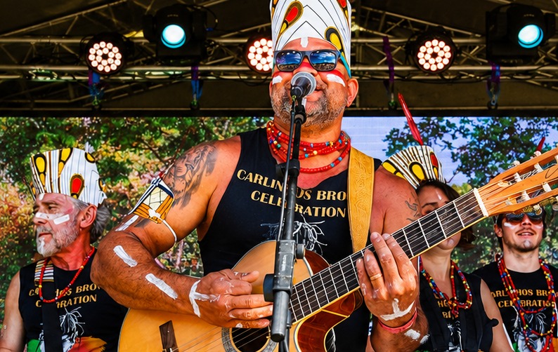 TRIBO musicians on stage in feathered headdresses, lead singer playing acoustic guitar into a microphone
