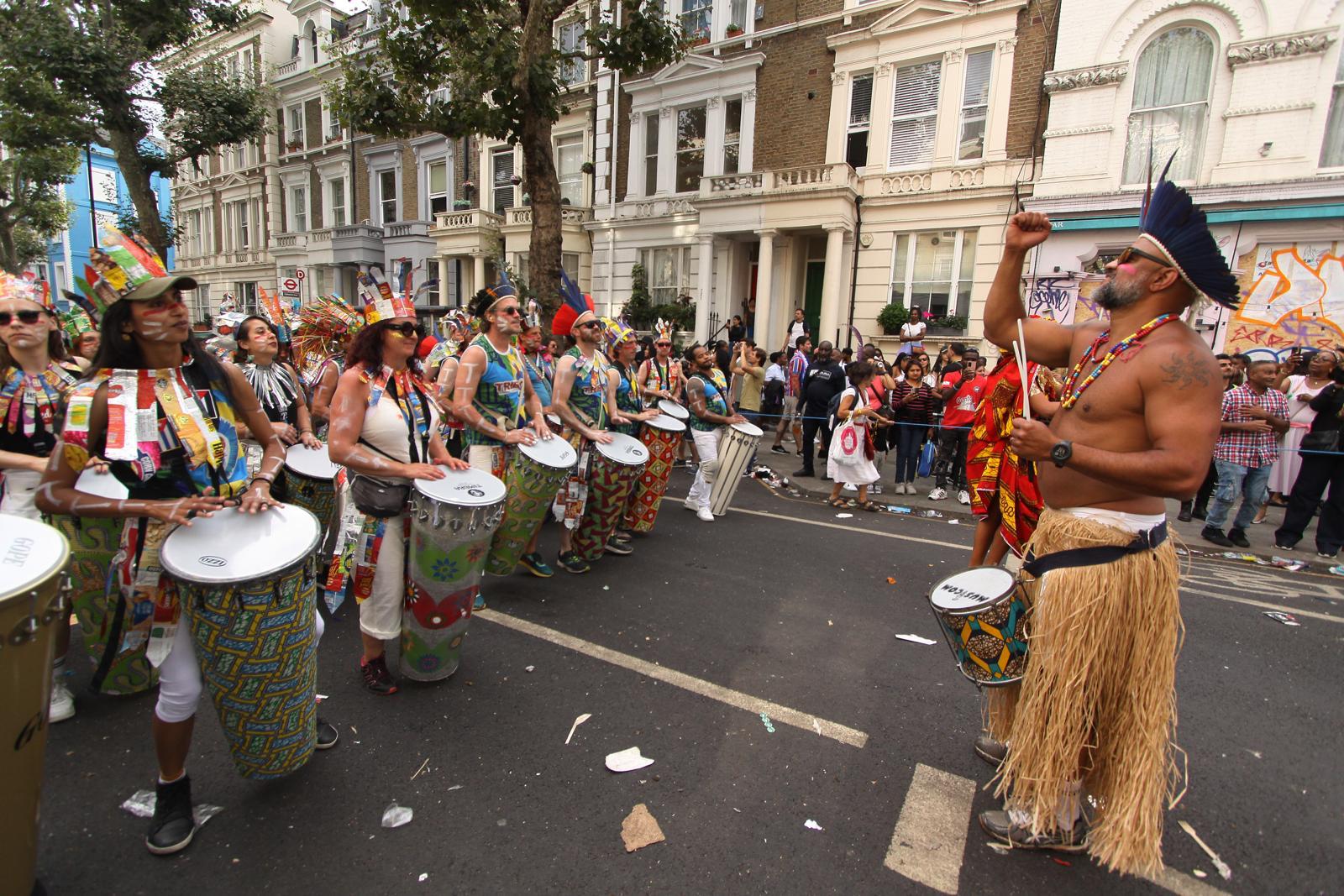 TRIBO percussionists in recycled costumes parading past white stucco houses at Notting Hill Carnival