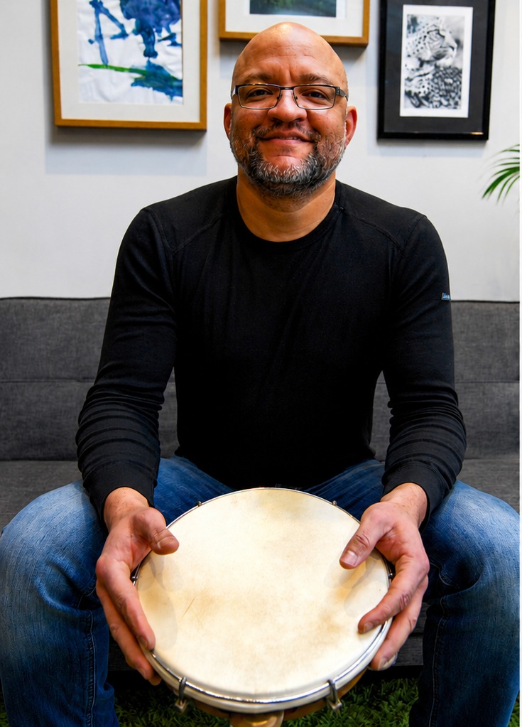 Marcos Santana seated holding a pandeiro, smiling to camera