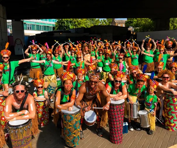 Full TRIBO bloco in green shirts and feathered headdresses raising drumsticks together