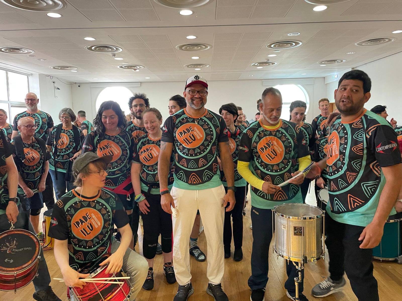 TRIBO members and workshop participants in Ipadê Ilú Percussion Festival shirts inside a bright hall, drums in hand